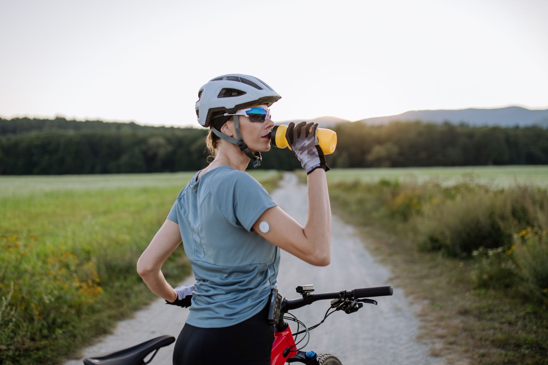 Diabetische Radfahrerin mit einem kontinuierlichen Blutzuckermessgerät am Arm trinkt Wasser während ihrer Radtour